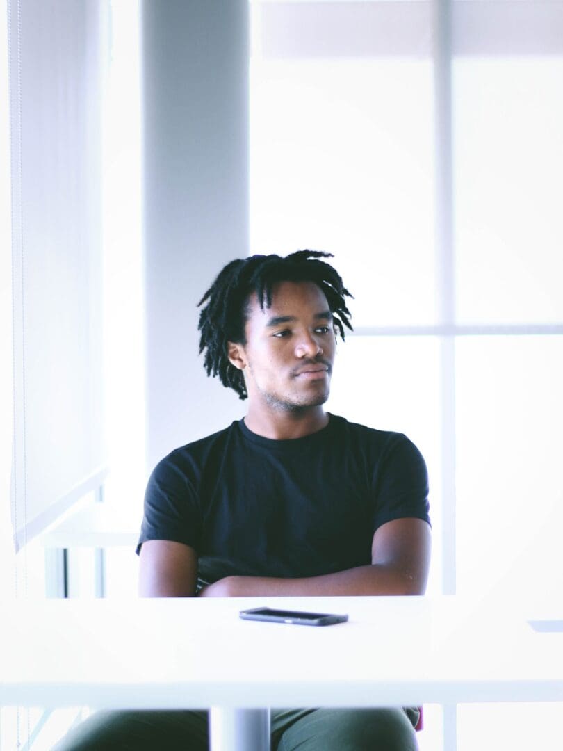 Black man sitting alone, thinking in a black shirt, contemplating the emotional effects of people-pleasing.